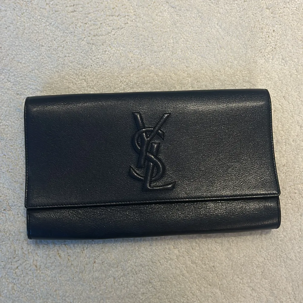 Elegant svart clutch från YSL i skinn med det ikoniska YSL-logotypen på framsidan. . Laukut & Käsilaukut.