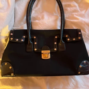Svart vintage handväska med nitar - Snygg svart handväska med nitar och ett stilrent spänne framtill. Väskan har två handtag och dekorativa detaljer i hörnen. Perfekt för att ge din outfit en edgy touch.