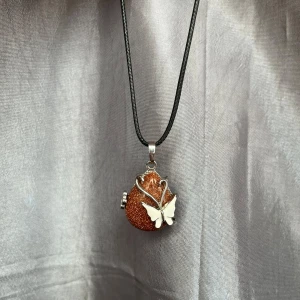 Halsband med glittrig brun sten och fjäril - Unikt halsband med en glittrig brun/orange solsten dekorerad med en vit och silverfärgad fjäril. Hänget hänger på en svart rem och har en elegant design. Aldrig använd. Går att byta kedja. Kommer inte ihåg exakt vart halsbandet är ifrån men tror jag köpt den i Dubai. 😊