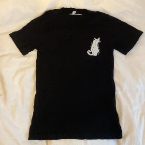 Svart t-shirt med kattmotiv från The Spark Company - Svart t-shirt i 100% bomull från The Spark Company. På bröstet finns ett tryck av en katt med stjärnor och månar samt texten 'Know Your Power'. Klassisk passform och korta ärmar. Perfekt för dig som gillar svarta basic tröjor o katter 🫶aldrig använd