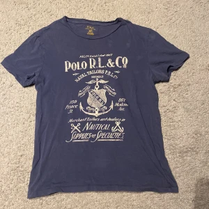 Blå t-shirt från Polo Ralph Lauren - Snygg blå t-shirt från Polo Ralph Lauren med vintageinspirerat tryck i vitt på framsidan. Modellen är normal passform och har korta ärmar. Perfekt för en avslappnad och stilren look. Väldigt ovanlig modell