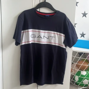 Svart t-shirt med logga från GANT - Snygg svart t-shirt från GANT med vit panel och röd detalj över bröstet samt stor GANT-logga framtill. Klassisk passform och rund halsringning. Perfekt för en stilren och avslappnad look.
