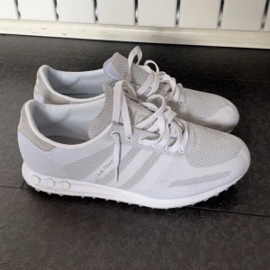 Vita Adidas LA Trainer sneakers - Säljer ett par fräscha vita Adidas LA Trainer sneakers med klassiska tre ränder på sidan. Skorna har snörning, rund tå och är tillverkade i syntetmaterial och mesh för en sportig look. Perfekta för dig som gillar stilrena och bekväma sneakers.