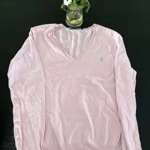 Ljusrosa v-ringad tröja från Ralph Lauren - Säljer en ljusrosa v-ringad tröja från Ralph Lauren med broderad logga på bröstet. Tröjan har långa ärmar och är tillverkad i bomull, vilket gör den mjuk och skön att bära. Perfekt för en stilren och klassisk look.