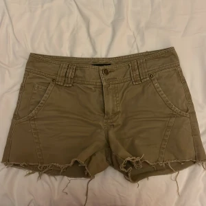 Beiga shorts från Massimo Dutti - Snygga beiga shorts från Massimo Dutti med fransig kant nertill och klassisk femficksdesign. Shortsen har bälteshällor och knappstängning framtill. Perfekta för varma dagar och enkel att matcha med olika toppar. Midjemått:39cm innerbenslängd: 7cm 