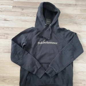 Svart hoodie från Peak Performance - Säljer en svart hoodie från Peak Performance med vit logga på bröstet. Klassisk modell med huva och känguruficka framtill. Perfekt för en avslappnad stil och enkel att matcha med det mesta.