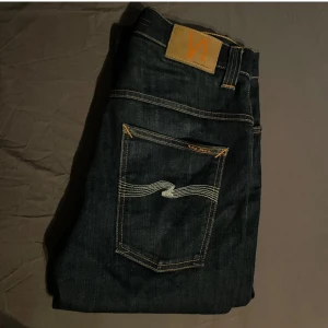 Mörkblå jeans från Nudie Jeans W31 L34 - Snygga mörkblå jeans från Nudie Jeans med klassiska orangea sömmar och bakficka med vågig detalj. Modellen har rak passform och normal midja. Perfekta för dig som gillar stilrena och tidlösa jeans.