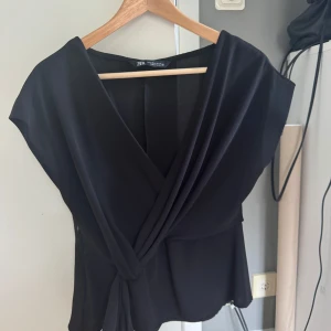 Svart omlottopp från Zara - Snygg svart omlottopp från Zara med korta ärmar. Toppen har en elegant omlottdesign framtill som ger en stilren look. Perfekt för en chic och modern stil.