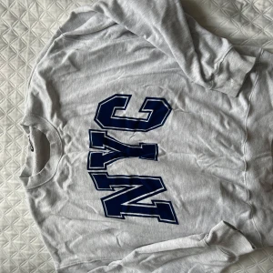 Grå sweatshirt med NYC-tryck - Säljer en grå sweatshirt med stort blått NYC-tryck på framsidan. Tröjan har långa ärmar och ribbade muddar vid ärmslut och nederkant. Perfekt för en avslappnad stil. Säljer även ett sett av mjukisbyxor + matchande hoodie som även passar till denna tröjan då dom är samma färg. Perfekt för att mixa till två olika outfits!🤩