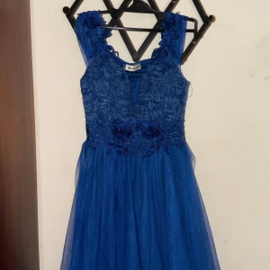 Blå spetsklänning med volangdetaljer - Elegant blå klänning med spetsöverdel och volangdetaljer. Klänningen har en v-ringad design och är ärmlös, vilket ger en stilren look. Perfekt för festliga tillfällen.