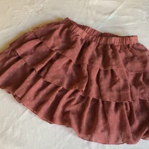 Rosa volangkjol från Shein - Charmig rosa kortkjol från Shein med flera lager av volanger och ett diskret prickigt mönster. Kjolen har en elastisk midja för bekväm passform. Perfekt för en somrig look!
