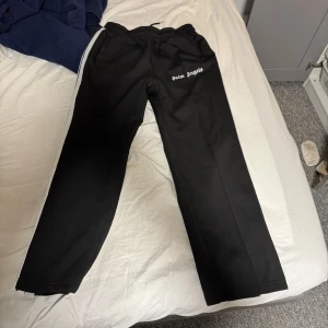 Svarta byxor från Palm Angels - Snygga svarta byxor från Palm Angels med vit text på ena benet. Byxorna har en normal passform och vita ränder längs sidorna. Perfekta för en stilren look.