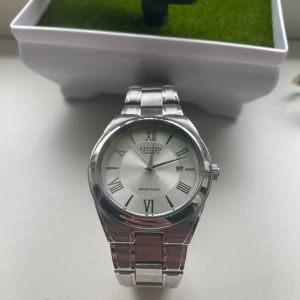 Silverfärgad klocka från Citizen Quartz - Stilren klocka från Citizen Quartz med silverfärgat armband i metall och vit urtavla. Klockan har romerska siffror, datumvisning och klassisk design. Perfekt accessoar för dig som gillar tidlös stil.