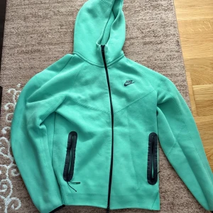 Turkos hoodiejacka från Nike - Säljer en turkos hoodiejacka från Nike med svart dragkedja och svarta detaljer vid fickorna. Jackan har huva och lång ärm, perfekt för träning eller chill. Nike-logga på bröstet och två fickor framtill.