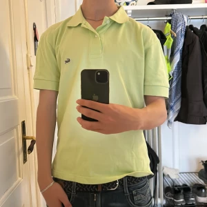 Ljusgrön piké t-shirt från Lacoste - Snygg ljusgrön piké t-shirt från Lacoste med klassisk krage och knappar framtill. Ikoniska krokodilloggan broderad på bröstet. Perfekt för en stilren och avslappnad look. Nypris: 1000kr Storlek: S/M