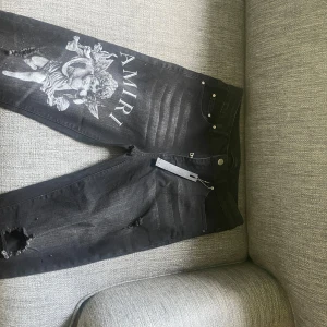 Svarta jeans med tryck från Amiri - Snygga svarta jeans från Amiri med ett unikt tryck av en ängel på ena benet. Jeansen har slitna detaljer och en klassisk femficksdesign. Perfekta för dig som vill ha en fet stil.