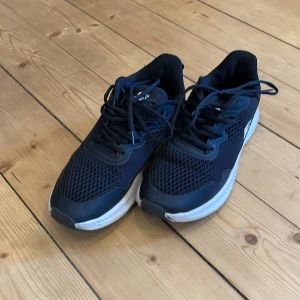 Svarta sneakers med vit sula - Säljer ett par svarta sneakers med vit sula och snörning. Skorna har en sportig design med meshdetaljer för extra andningsförmåga. Perfekta för träning eller vardagsbruk.