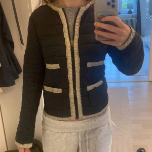 Vår/höst jacka - Säljer en svart quiltad jacka från Mango med beige volangdetaljer längs dragkedjan, kragen och fickorna. Jackan har fyra fickor framtill och stängs med dragkedja. Perfekt för dig som gillar en stilren look med twist.