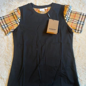 Svart t-shirt med rutiga ärmar från Burberry - Säljer en svart t-shirt från Burberry med klassiska beige-rutiga ärmar och detaljer vid halsen. T-shirten har rund halsringning och är kortärmad. Ikonisk design som sticker ut med Burberrys signaturrutor på ärmarna. Jag är 178 och väger 66 den sitter precis på mig (lite tajt) KVITTO MEDFÖLJER!