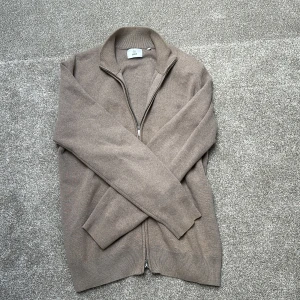 Beige stickad kofta med dragkedja - Säljer en stilren beige stickad kofta från Uniqlo med dragkedja framtill och hög krage. Perfekt för lager-på-lager och passar till många olika outfits. Klassisk och enkel design med ribbade muddar.