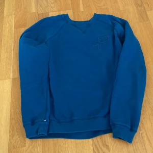Blå sweatshirt från Pelle P, Stl S - Säljer en blå sweatshirt från Pelle P med broderad logga på bröstet. Tröjan har rund halsringning, långa ärmar och ribbade muddar vid ärmslut och nederkant. Herr storlek S