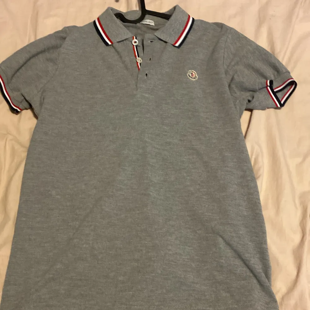 Säljer en grå piké t-shirt från Moncler med klassisk krage och knappar framtill. Kragen och ärmsluten har röd, vit och marinblå randig detalj. Liten Moncler-logga broderad på bröstet. Perfekt för en stilren look.. T-paidat.