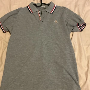 Grå piké t-shirt från Moncler - Säljer en grå piké t-shirt från Moncler med klassisk krage och knappar framtill. Kragen och ärmsluten har röd, vit och marinblå randig detalj. Liten Moncler-logga broderad på bröstet. Perfekt för en stilren look.