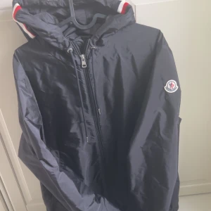 Mörkblå vindjacka från Moncler - Snygg mörkblå vindjacka från Moncler med huva och dragkedja framtill. Jackan har Moncler-logga på ärmen och vita samt röda detaljer på huvan. Perfekt för blåsiga dagar.