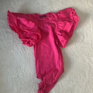Rosa body med volangärmar - Säljer en snygg rosa body med stora volangärmar. Perfekt för att göra ett statement! Den har en rund halsringning och knäppning i grenen för enkel på- och avtagning.