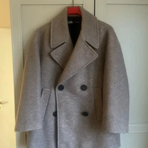 Beige kappa från Zara - Snygg beige kappa från Zara med dubbelknäppning och breda slag. Tillverkad i ett ulligt material som ger en lyxig känsla. Perfekt för kyligare dagar.