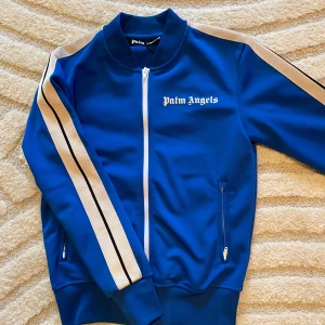 Palm Angels Tröja - Säljer en snygg blå track jacket från Palm Angels med vita detaljer och dragkedja. Jackan har långa ärmar med beige och vita ränder längs sidorna. Perfekt för en sportig look eller avslappnad stil. Storlek XS. Använd några gånger, jätte fint skick. Ny pris 3500. Prislapp finns kvar.