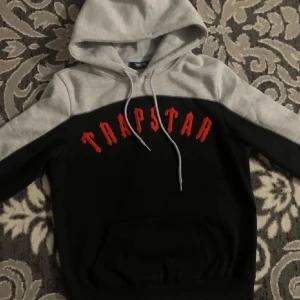 Svart och grå hoodie från Trapstar - Snygg hoodie från Trapstar i svart och grått med röd text på bröstet. Den har en klassisk känguruficka och justerbar huva med snören. Perfekt för en avslappnad stil.