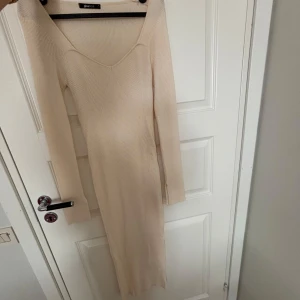 Beige ribbad klänning från Gina Tricot - Snygg beige långklänning från Gina Tricot i ribbat material. Klänningen har en v-ringad design och långa ärmar, perfekt för en stilren look. Passar utmärkt till både vardag och festliga tillfällen.