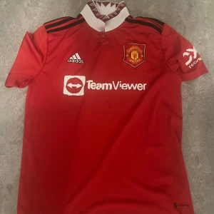 Röd fotbollströja från Adidas - Säljer en röd fotbollströja från Adidas med Manchester Uniteds emblem på bröstet. Tröjan har korta ärmar och vita detaljer, inklusive logotypen för TeamViewer. Perfekt för fans av laget!
