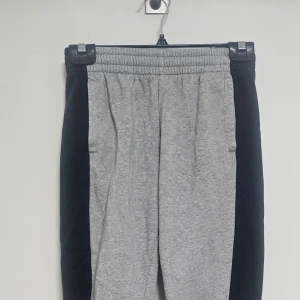 Grå mjukisbyxor från Adidas med svarta detaljer - Säljer ett par grå mjukisbyxor från Adidas med svarta paneler längs sidorna och stort Adidas-tryck på vänster ben. Byxorna har resår i midjan och mudd vid bensluten. Perfekta för chill eller träning.