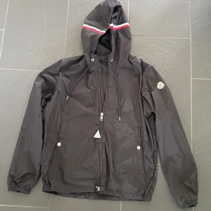 Svart vindjacka från Moncler med huva - Svart vindjacka från Moncler med huva och dragsko. Jackan har en diskret logotyp på ärmen och en dragkedja framtill. Huvan har en röd och vit rand som detalj. Två fickor med knappar på sidorna och elastiska muddar vid ärmslut.