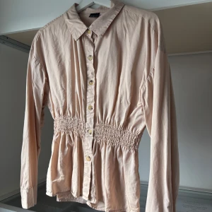 Beige blus med in dragen midja  - Säljer en beige blus från Gina tricot  med smockad midja och långa ärmar. Blusen har klassisk krage och knappar framtill. Perfekt för dig som gillar en feminin och avslappnad stil. Materialet känns mjukt och lätt.