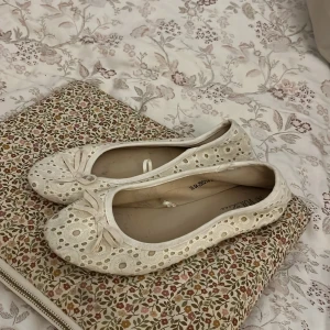 Vita ballerinaskor med spets och rosett - Säljer ett par vita ballerinaskor från Sthlm DG med spetsdetaljer och små rosetter framtill. Skorna har rund tå och är täckta av ett blommigt hålmönster som ger en romantisk känsla.