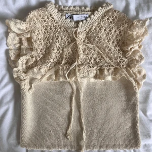 Beige spets- och stickad topp från Zara - Supersöt beige topp från Zara med spetsdetaljer och volanger på axlarna. Överdelen är i spets med knyt framtill och underdelen är stickad. 