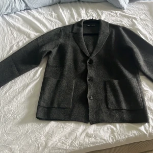 Mörkgrå kavaj från Zara - Säljer en stilren mörkgrå kavaj från Zara med klassisk krage och tre knappar framtill. Kavajen har två stora fickor och är långärmad, perfekt för en dressad look.