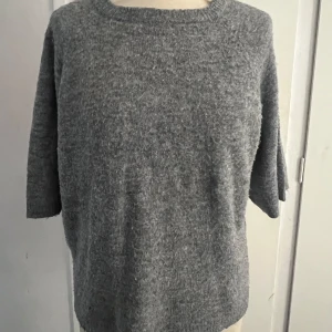 Grå stickad tröja från 157 - Mysig grå stickad tröja från 157 i storlek XS. Tröjan har kort ärm och rund halsringning, perfekt för våren. Enkel och stilren design som passar till det mesta.