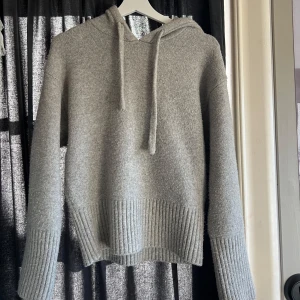 Grå stickad hoodie - Mysig grå stickad hoodie med huva och dragsnören. Tröjan har långa ärmar och bred ribbstickning vid ärmslut och nederkant. Perfekt för kyliga dagar och enkel att matcha med det mesta.