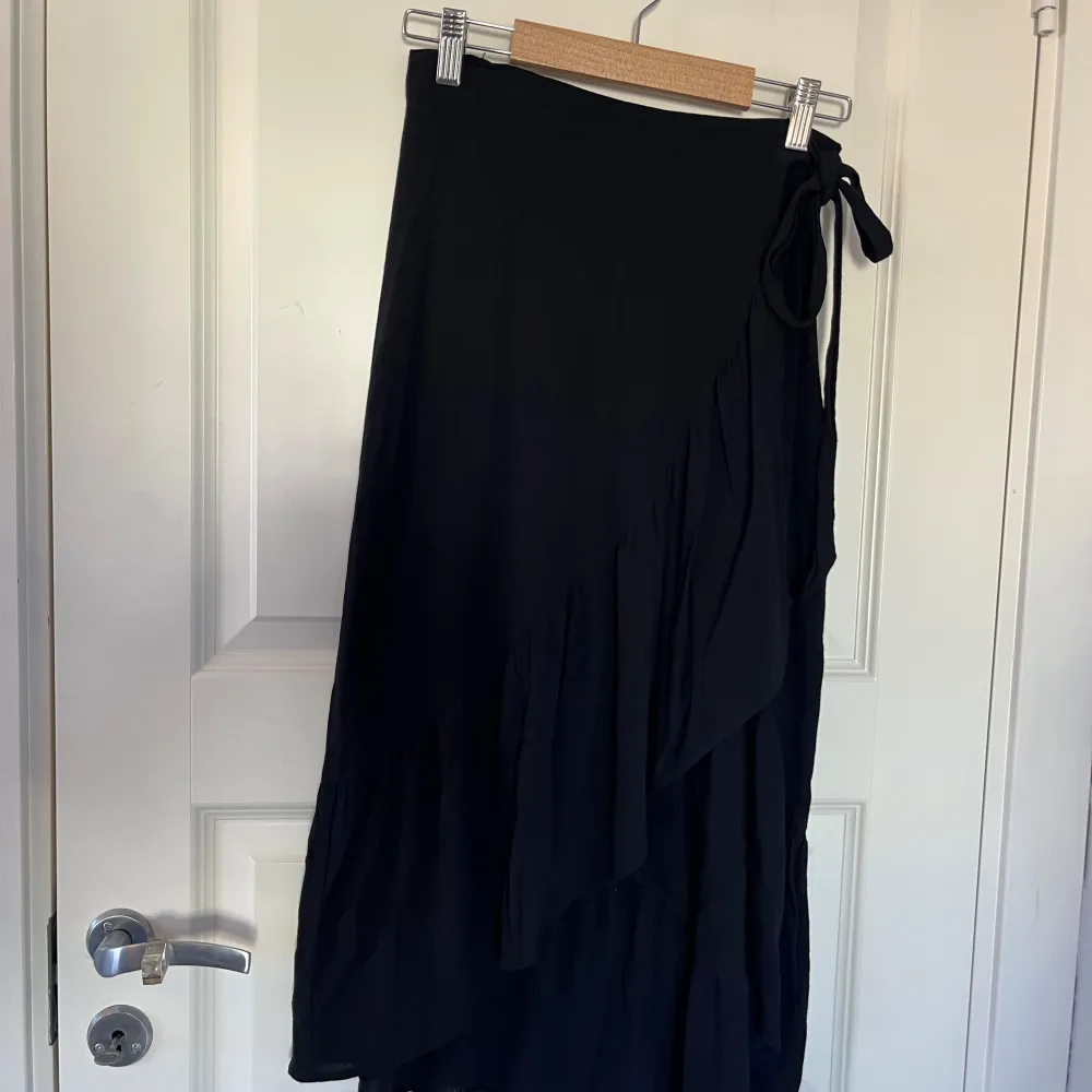 Säljer en svart lång omlottkjol med volangdetaljer och knytband i midjan. Kjolen har en elegant och flowy känsla, perfekt för dig som gillar stilrena plagg med lite extra detalj. Passar till många olika looks.. Hameet.