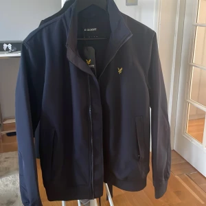 Mörkblå jacka från Lyle & Scott - Säljer en stilren mörkblå jacka från Lyle & Scott med dragkedja och klassisk gul logga på bröstet. Jackan har hög krage, två fickor med dragkedja och ribbade muddar. Perfekt för dig som vill ha en enkel och snygg look.