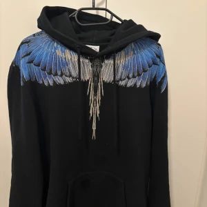 Svart hoodie från Marcelo Burlon - Säljer en svart hoodie från Marcelo Burlon med ett unikt blått och vitt vingmönster över axlar och bröst. Tröjan är endast använd vid ett fåtal tillfällen och är i ett extremt bra skick. Skriv gärna vid frågor😁