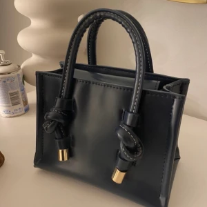 Svart handväska med knutdetaljer - Säljer en stilren svart handväska med två handtag och snygga knutdetaljer med guldfärgade avslut. Väskan är i slät syntet och har ett ljust innerfoder samt magnetlås. 