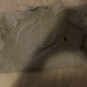 Grå piké t-shirt från Ralph Lauren - Säljer en klassisk grå piké t-shirt från Ralph Lauren med liten broderad logga på bröstet. T-shirten har korta ärmar och knappar vid kragen. Perfekt för en stilren och avslappnad look.