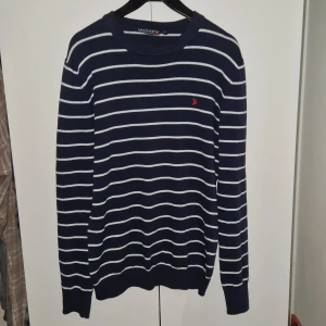 Mörkblå randig tröja från Gant - Snygg mörkblå tröja från Gant med vita ränder och en liten röd logga på bröstet. Tröjan har långa ärmar och ribbade muddar vid ärmslut och nederkant. Perfekt för en stilren look! Helt ny!