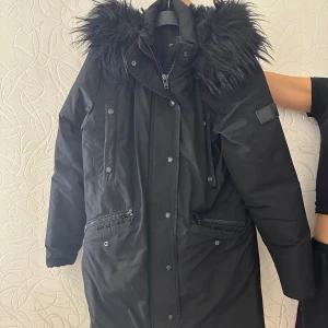 Svart parkas från Rockandblue med fuskpäls - Svart parkas från Rockandblue med stor huva och fluffig svart fuskpäls. Jackan har dragkedja och knappar framtill, flera fickor med dragkedja och tryckknappar. Perfekt för kalla dagar med sin långa modell och snygga detaljer.