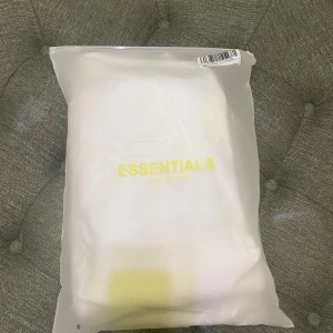 Vit Essentials Fear of God hoodie - Den är kopia1:1 Säljer en clean vit hoodie från Essentials Fear of God med svart logga på bröstet och ärmen. Klassisk känguruficka framtill och huva. Perfekt för dig som gillar streetwear och vill ha en stilren look.
